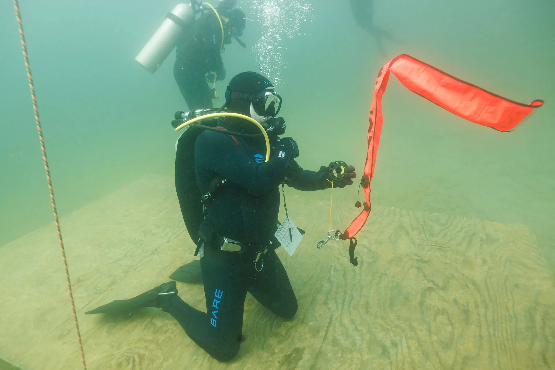 A SCUBA diver inflates a surface marker buoy while kneeling on a platform underwater