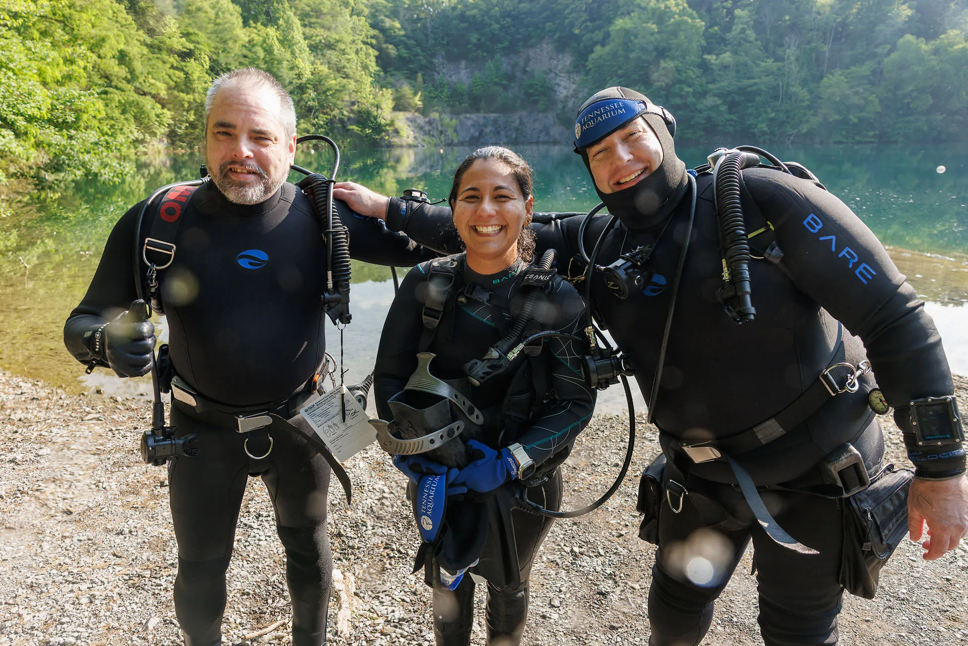 Three SCUBA divers pose for a photo on dry land near the shore of a quarry