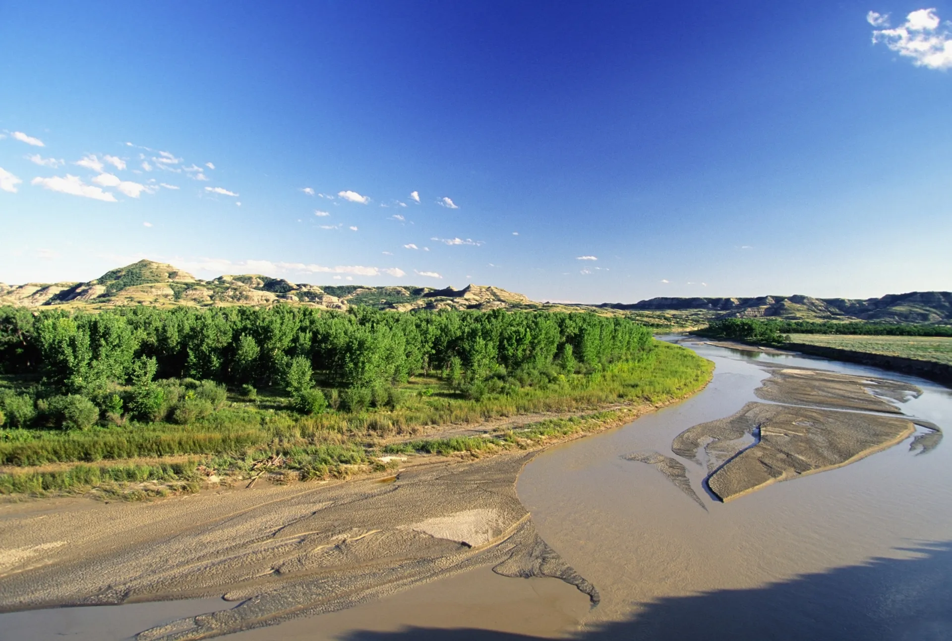 A high angle, aerial view of the Missouri River flowing through North Dakota. The river's water looks brown and muddy from massive amounts of sediment. The sky above is blue with only a few scattered high-altitude clouds.