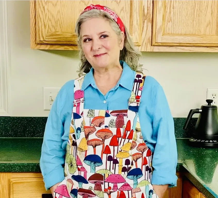 Sherri Castle standing in a kitchen with mushroom-patterned overalls on. Sherri is an award-winning professional food writer, recipe developer, cooking teacher and public speaker known for melding stories, humor and culinary expertise.